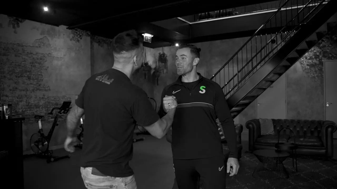 Still uit de film Wij zijn Sportlijf: twee mannen begroeten elkaar in een donkere sportschool